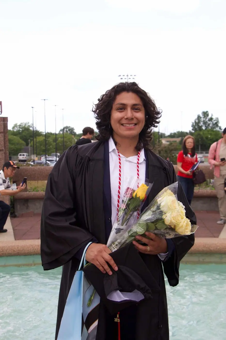 Bryan in cap and gown at his college graduation, holding bouquets beside a fountain