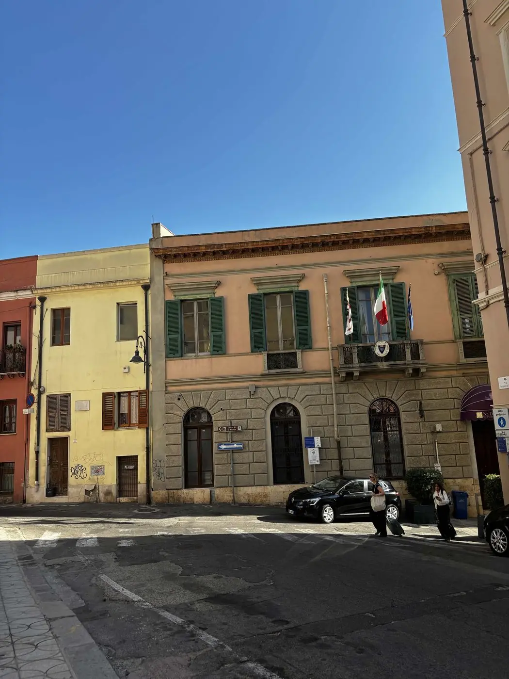 A sunlit Sardinian street of pastel buildings with green shutters and Italian flags